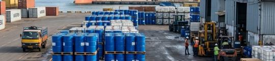 Illustration of hazard-labelled solvent drums and IBC totes stored in a shaded warehouse area in hot sunlight